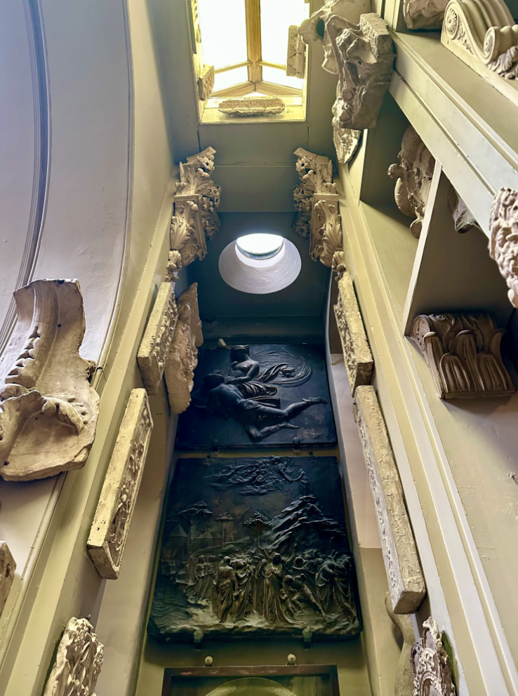 Museum room with small round skylight over antique architectural friezes, possibly bronze.