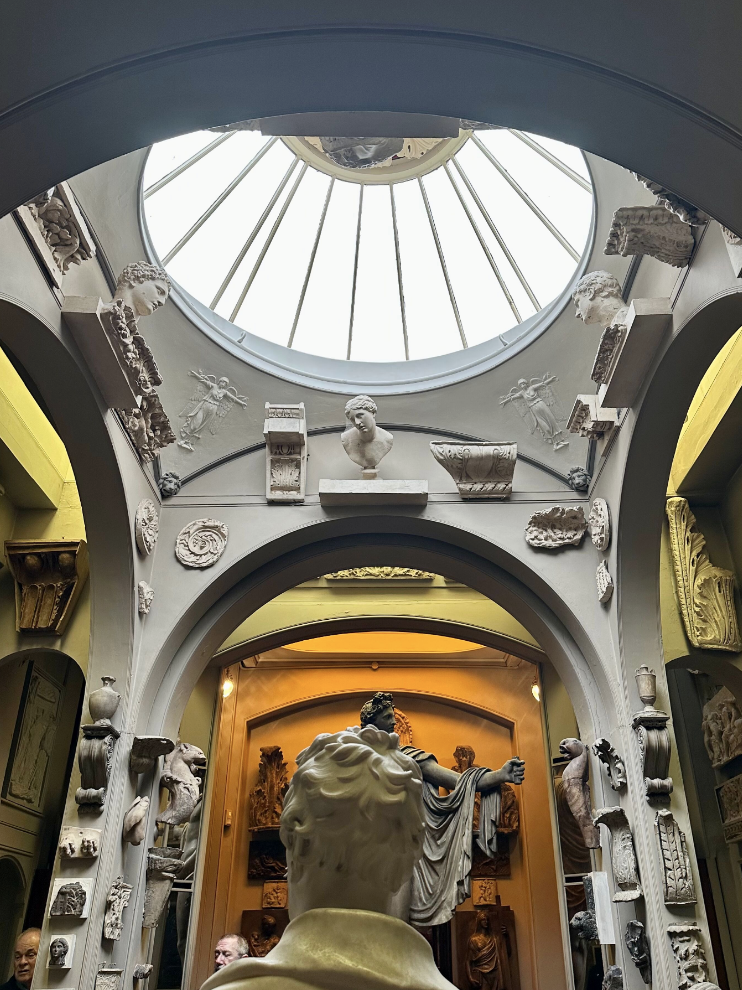 Museum room with domed skylight crowded with antiquities, statues, architectural elements.