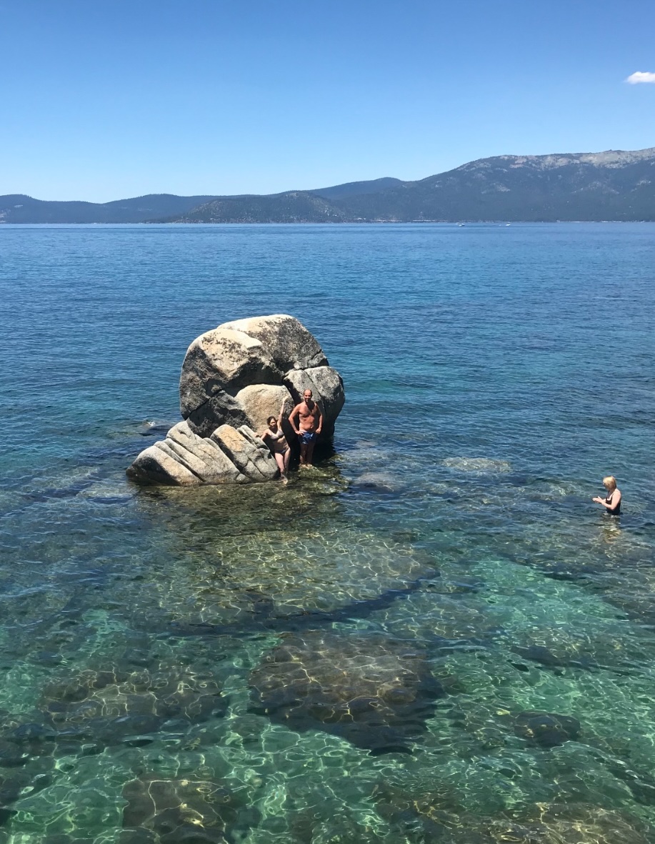 Three people swimming in a lake with a big rock.